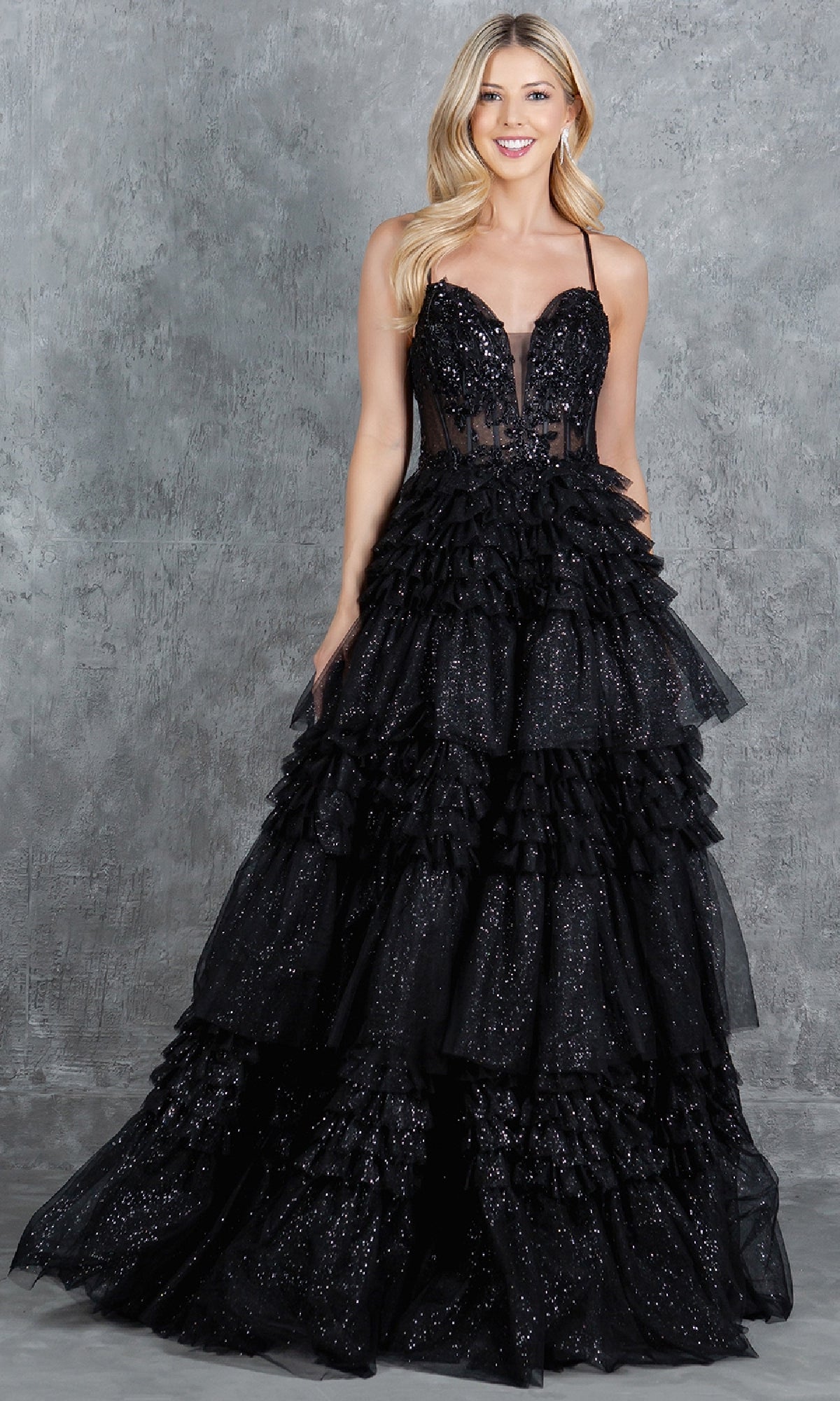 A woman in the Glitter Prom Ball Gown: Cinderella Couture 8163J—black, strapless, floor-length with ruffled layers and sparkling details—stands against a gray textured background.